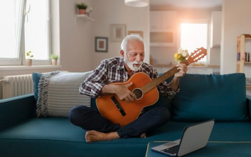 Happy good looking elderly man in a plaid shirt sits on a sofa in the living room and learns to play acoustic guitar online using a Laptop