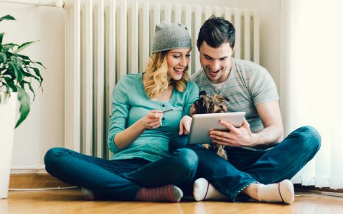 Young Couple Sitting On Floor in a living room at home and shopping online with digital tablet. Smiling woman with hat holding credit card, and man holding tablet and their cute dog on the lap.